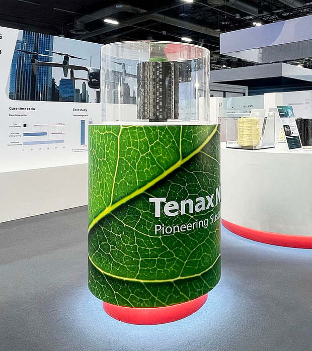 Ein runder Messestand mit einem großen grünen Blatt-Design und dem Logo "Tenax Pioneering Sus", der eine zylindrische Kohlefaserstruktur zeigt. Im Hintergrund sind Diagramme zu "Cure time ratio" und "Cost study" sichtbar.
