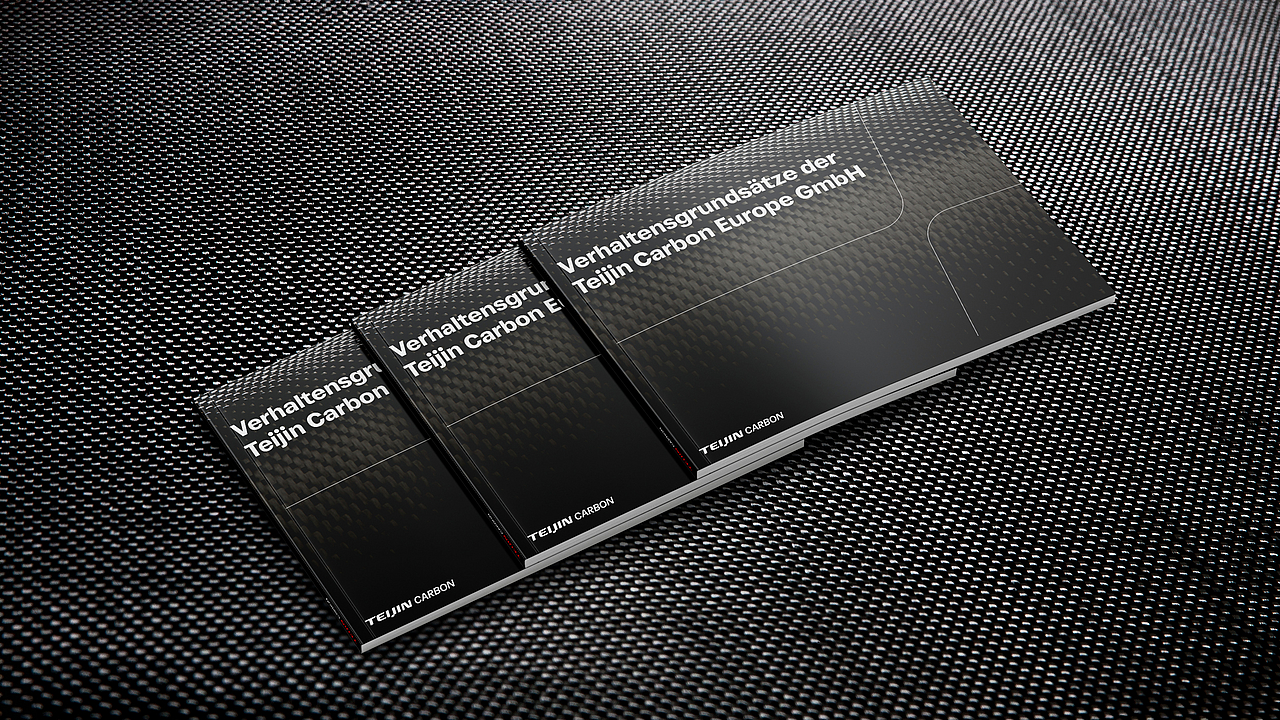 Drei gestapelte schwarze Broschüren mit der Aufschrift "Verhaltensgrundsätze der Teijin Carbon Europe GmbH" auf Carbonfaser-Hintergrund.