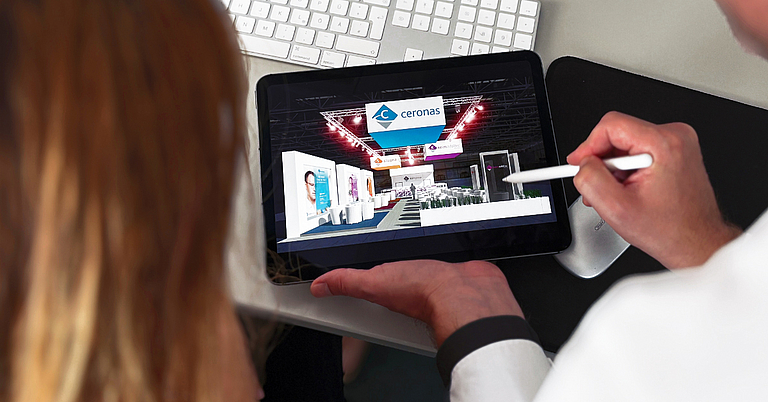 Zwei Personen betrachten auf einem Tablet die digitale Visualisierung eines Messestands. Eine Person zeigt mit einem Stift auf das Designkonzept des Messestands, während im Hintergrund Tastatur und Arbeitsplatz zu sehen sind. Die Szene zeigt die Planung und Konzeption eines Messeauftritts.