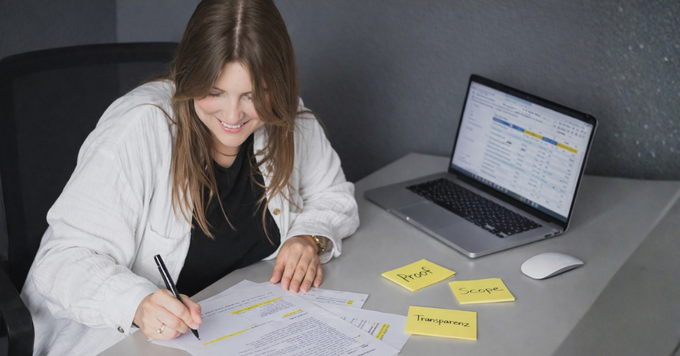 Eine Frau sitzt an ihrem Schreibtisch und prüft ausgedruckte Marketingtexte mit einem Stift. Auf dem Tisch liegen Notizzettel mit den Begriffen ‚Proof‘, ‚Scope‘ und ‚Transparenz‘, daneben ein Laptop mit Datenübersicht. Die Szene zeigt die kritische Prüfung von Nachhaltigkeitsaussagen im Marketing.