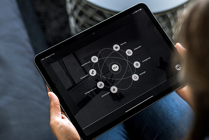 Eine Person hält ein Tablet in den Händen, auf dessen Bildschirm eine grafische Darstellung zum Thema SEA (Search Engine Advertising) mit Elementen wie Planung, Zielgruppe, Strategie, Anzeigen und Erfolgsmessung zu sehen ist.