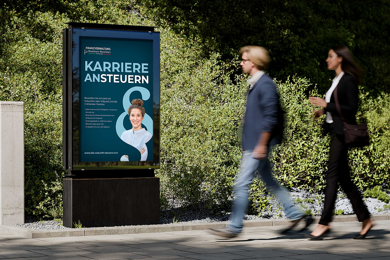 Zwei Personen gehen an einem Werbeplakat der Finanzverwaltung NRW mit der Aufschrift "Karriere ansteuern" vorbei.