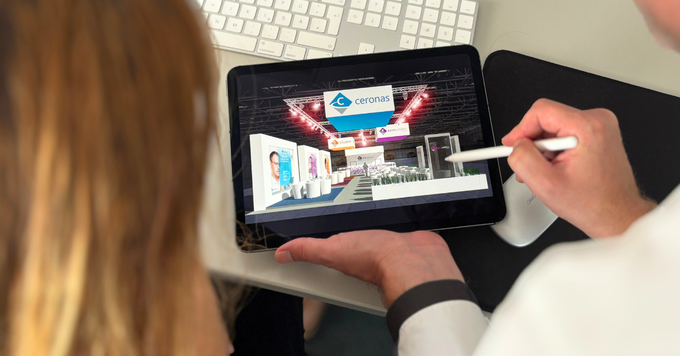 Zwei Personen betrachten auf einem Tablet die digitale Visualisierung eines Messestands. Eine Person zeigt mit einem Stift auf das Designkonzept des Messestands, während im Hintergrund Tastatur und Arbeitsplatz zu sehen sind. Die Szene zeigt die Planung und Konzeption eines Messeauftritts.