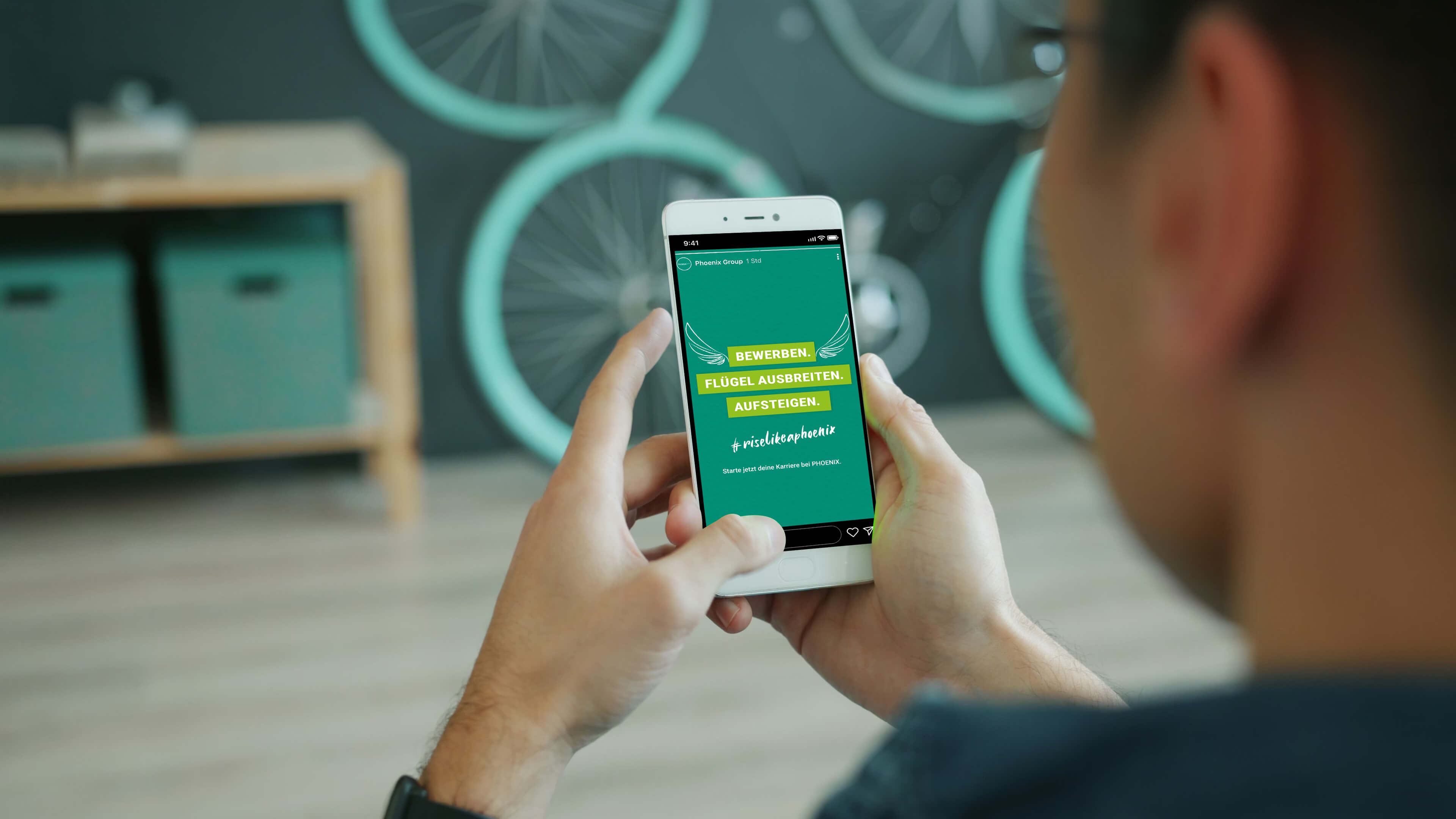 Person hält ein Smartphone mit der Phoenix Group Recruiting-Anzeige „Bewerben. Flügel ausbreiten. Aufsteigen.“ in den Händen.