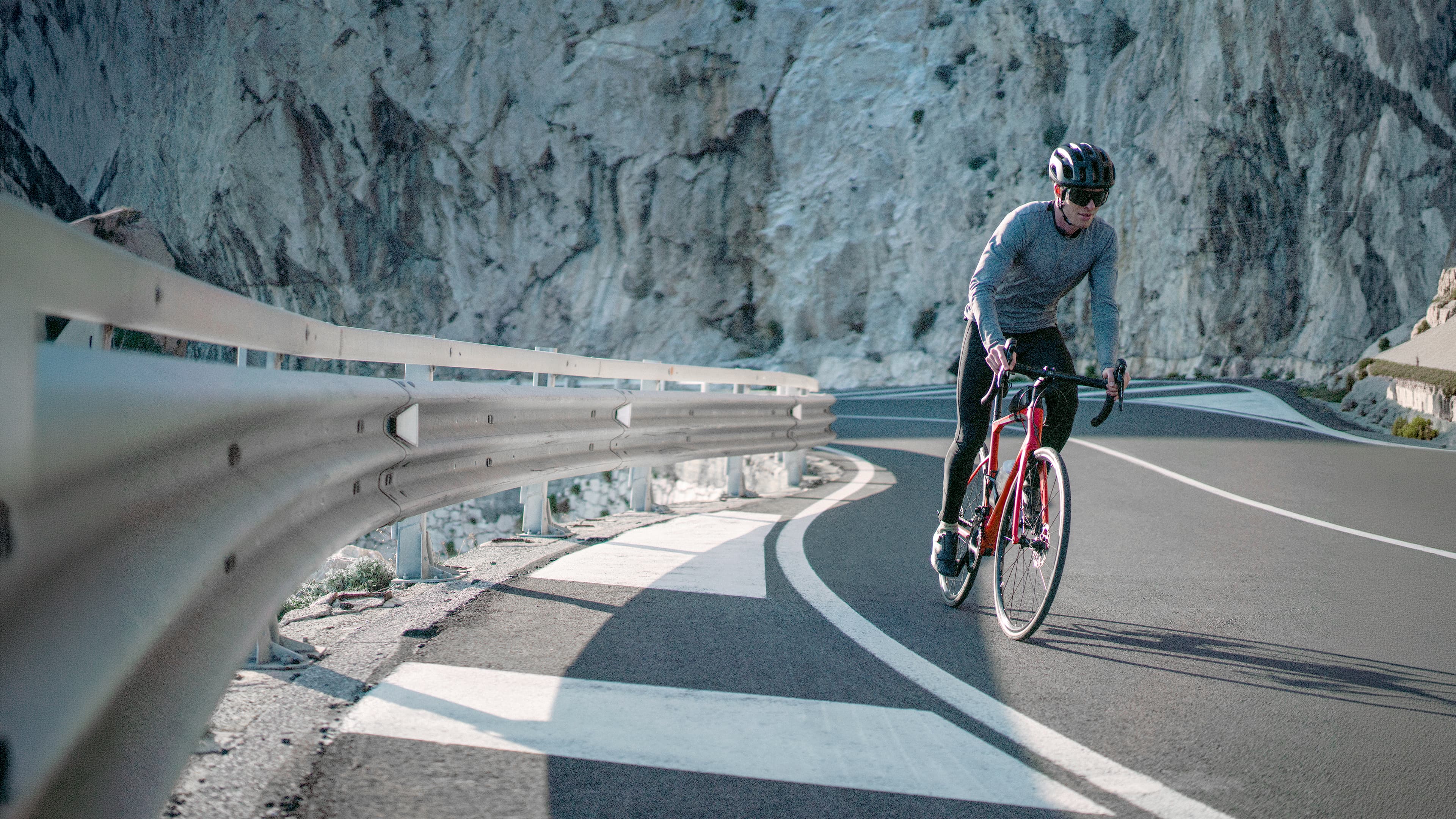 Radsportler mit Helm und Sportbrille fährt mit einem roten Rennrad durch eine asphaltierte Serpentine vor einer hellen Felswand.